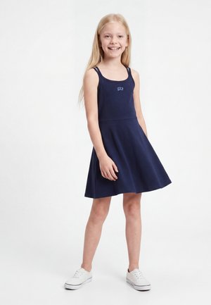 Νεαρό κορίτσι χαμογελά, στέκεται, φοράει ένα μπλε φόρεμα χωρίς μανίκια με το λογότυπο "GAP" και λευκά αθλητικά παπούτσια σε λευκό φόντο.