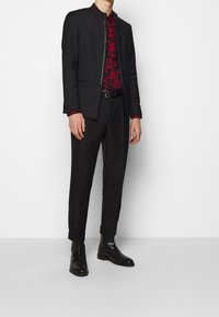 Schwarzer maßgeschneiderter Anzug mit Stehkragen, darunter ein rot gemustertes Hemd. Ergänzt durch schwarze slim-fit Hosen und Ankle Boots.