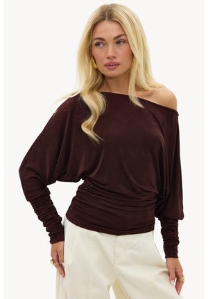 Femme portant un haut long à manches longues marron foncé, épaules dénudées avec manches froncées, et un pantalon crème taille haute, regardant de côté.