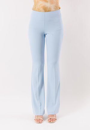 Donna che indossa pantaloni a zampa color azzurro chiaro aderenti e sandali beige con punta aperta, in piedi contro uno sfondo bianco neutro.