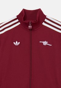 Burgundská zipsová bunda s vysokým golierom; obsahuje biele a zlaté pruhy a vyšívané logá Adidas a delá.
