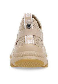 Beige sportskor med en texturerad stickad överdel, ett plastband över mellanfoten och en distinkt hälklack med vita varumärkesdetaljer.