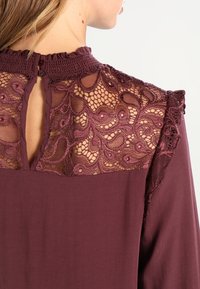 Abito borgogna caratterizzato da dettagli in pizzo intricati sulla schiena, con chiusura a bottone e un tessuto morbido per una vestibilità fluida.