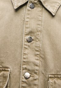 Κοντινό πλάνο στο μπροστινό μέρος μπεζ τζιν μπουφάν με μεταλλικά κουμπιά χαραγμένα με "Mads Nørgaard", γιακά και μερικές τσέπες ορατές.