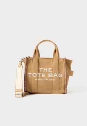 Καφέ τσάντα tote από καμβά με διπλές λαβές στο πάνω μέρος, αποσπώμενο ιμάντα ώμου, λευκό κείμενο "THE TOTE BAG MARC JACOBS" και δομημένο σχέδιο.