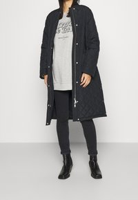 Cappotto lungo nero imbottito con bottoni a pressione e zip, indossato sopra una felpa grigia e jeans neri, abbinato a stivaletti neri.