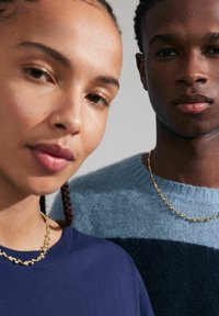 Zwei Modelle tragen einen dunkelblauen Sweatshirt und einen hellblauen Pullover mit dunkelblauen Streifen. Beide tragen goldene Halsketten.