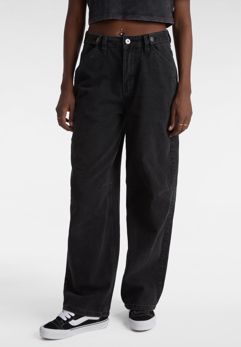 Personne portant un jean noir ample, un crop top noir et des baskets noires et blanches, debout avec les bras détendus le long du corps devant un fond uni.