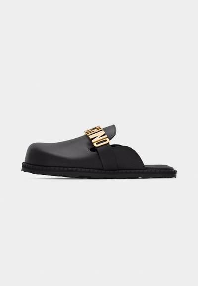 Svart slip-on-träskosko med öppen häl och guldig "HINO"-text på rem över ovansidan, platt gummisula, sidvy på vit bakgrund.