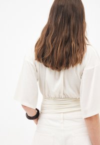 Mujer con cabello largo y castaño que lleva una blusa blanca suelta de mangas anchas y pantalones blancos de tiro alto, vista desde atrás.