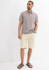 Polo grigio, pantaloni shorts beige chiaro con risvolti e sandali scuri. Il modello è in piedi su uno sfondo bianco, mostrando un abbigliamento estivo casual.