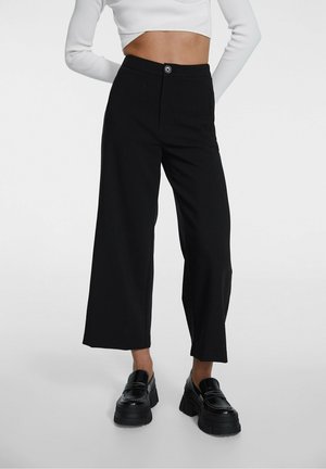 Vrouw met zwarte wijde cropped broek, wit cropped top met lange mouwen en zwarte chunky platform loafers, staat met handen achter de rug.