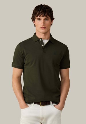 Joven con cabello castaño rizado que lleva una camisa polo de manga corta verde oscuro y pantalones blancos con las manos en los bolsillos.