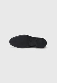 Suela de un zapato de vestir negro de cuero con patrones de agarre texturizados y la marca "Tommy Hilfiger", presentada sobre un fondo blanco liso.