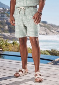Pantaloni corti verde chiaro con motivo, realizzati in tessuto testurizzato, con vita elastica e cordoncino. Indossati con sandali beige su un ponte in legno.