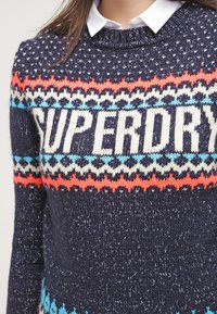 Laivastonsininen neulepaita, jossa on näyttävä valkoinen "SUPERDRY"-logo ja monivärisiä geometrisiä kuvioita oranssina, sinisenä ja kermaisena rinnassa.