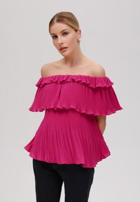 Avtale-bluse med avtagbare ermer i lysende rosa, i plissert stoff med lagdelte volanger. Myk tekstur og flytende design, kombinert med svarte bukser.