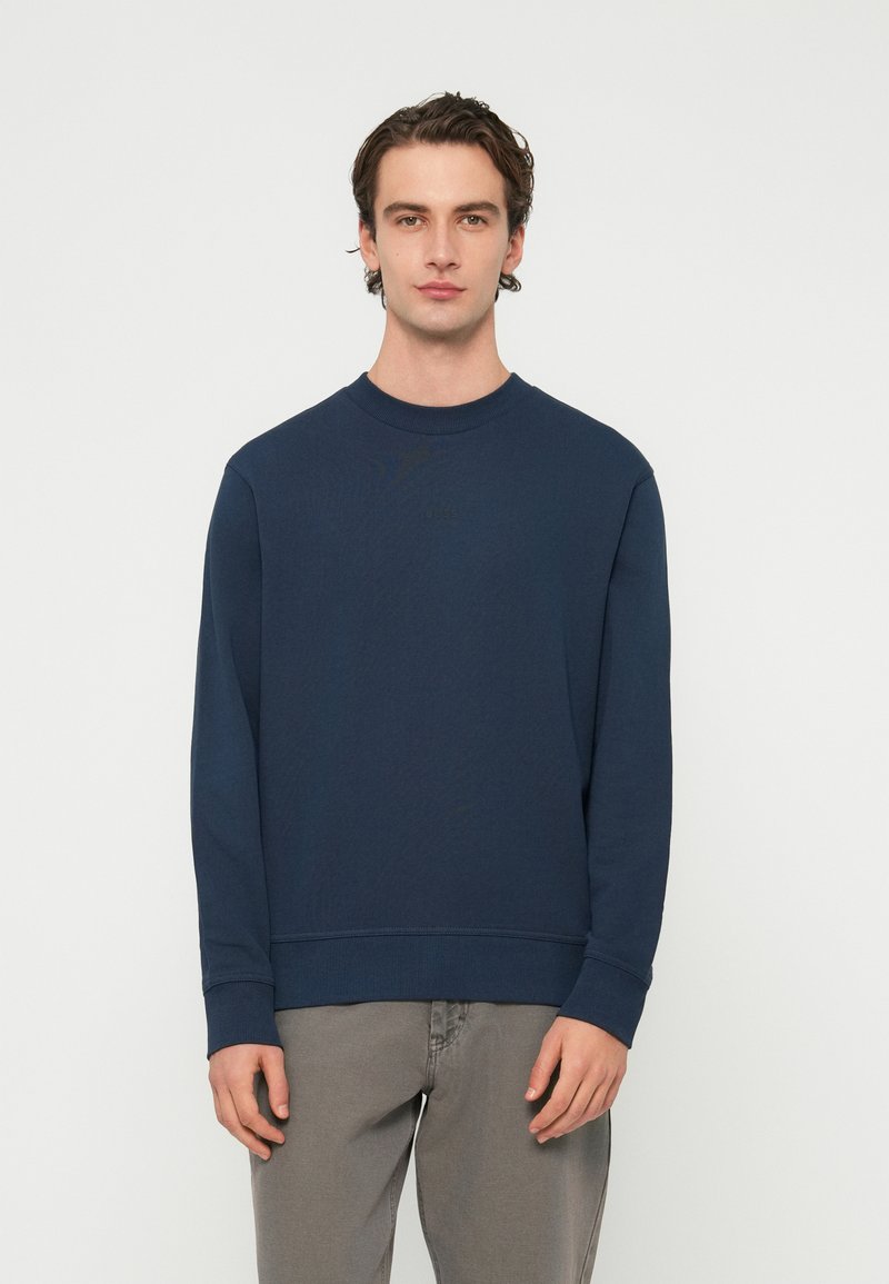 Mladý muž v obyčajnej tmavomodrej mikine a svetlosivých nohaviciach stojaci na bielom pozadí.