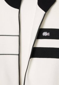 Veste blanche avec fermeture éclair noire, col côtelé noir, rayures horizontales noires et petit logo crocodile brodé sur la poitrine.