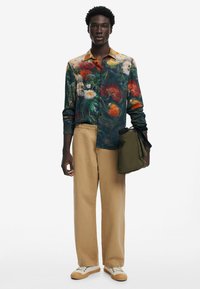 Camicia button-up con motivo floreale di colore verde scuro, con accenti gialli, rossi e bianchi. Pantaloni beige a gamba dritta e una borsa verde scuro. Sneakers bianche.