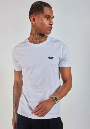T-shirt en coton blanc avec un col rond, des manches courtes et un logo noir sur la poitrine gauche ; porté avec un short noir et une montre argentée.