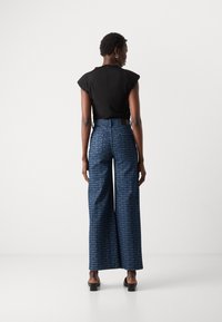 Schwarze Bluse mit kurzen Ärmeln und Reißverschluss auf der Rückseite, kombiniert mit hochtaillierten, weit geschnittenen blauen Hosen mit wiederholtem weißen Logo-Muster.