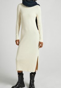 Stickad, ribbad klänning i krämfärg med långa ärmar och sidofickor. Bärs med höga svarta stövlar och en texturerad mörk scarf runt halsen.