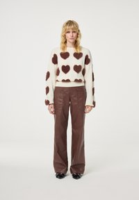 Crème trui met bruine fuzzy hartpatronen, gecombineerd met bruine leren wijde broek. Model staat op een effen witte achtergrond.