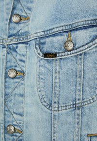 Svetlo modra denim jakna s srebrnimi kovinskimi gumbi, vidno šivanje in dvema žepoma na prsih, ki imata črne in rumene oznake Lee.