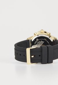 Correa de reloj de goma negra con hebilla de metal dorado, que presenta una caja redonda en tono dorado y una esfera negra con marcadores y botones visibles.