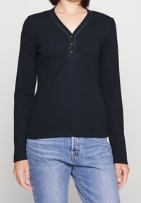 Czarna bluzka z długim rękawem, dekoltem w serek i zapięciem na trzy guziki, o prążkowanej fakturze i dopasowanym kroju. W zestawie z jasnoniebieskimi dżinsami.