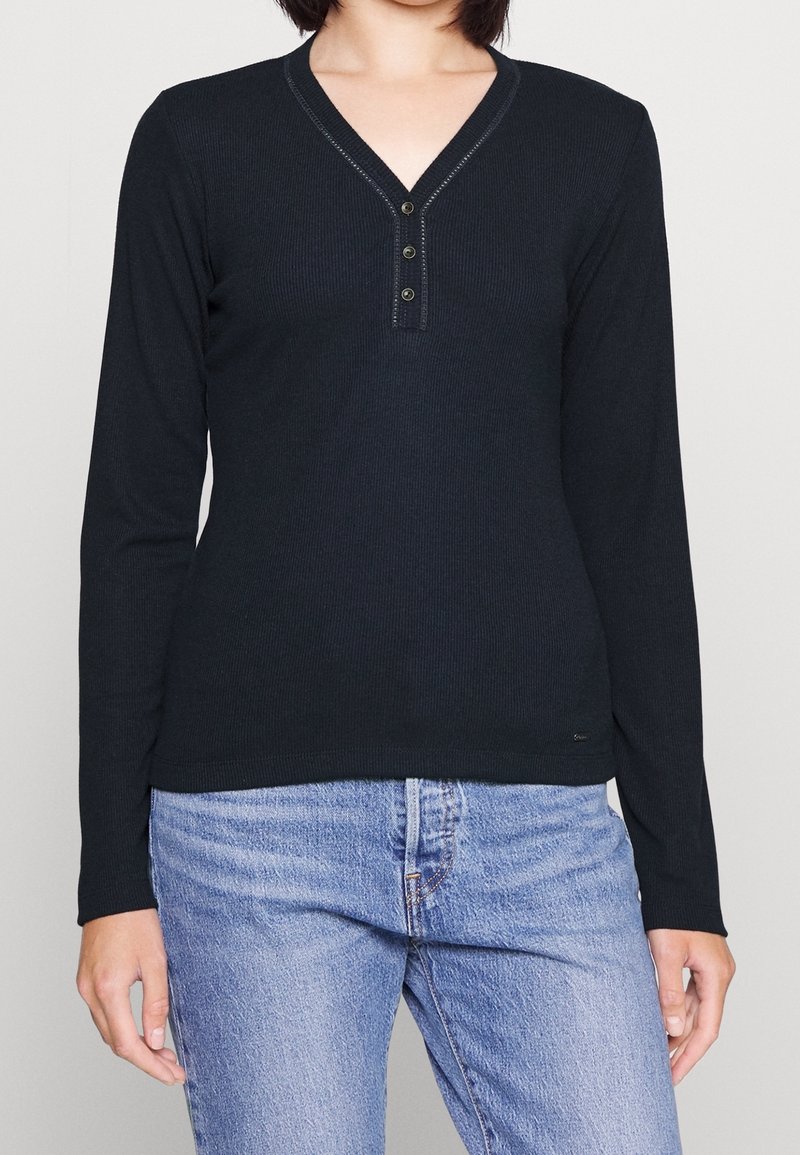 Czarna bluzka z długim rękawem, dekoltem w serek i zapięciem na trzy guziki, o prążkowanej fakturze i dopasowanym kroju. W zestawie z jasnoniebieskimi dżinsami.