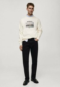 Krem sweatshirt z natisnjeno podobo mestne silhuete z besedilom "VELIKI JABOLK". V kombinaciji z črnimi hlačami in črnimi čevlji.