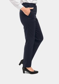 Marineblauwe broek met een tailor fit, voorzien van zijzakken en een elastische tailleband. Gedragen met zwarte gesloten teenschoenen, wat een strakke silhouet benadrukt.