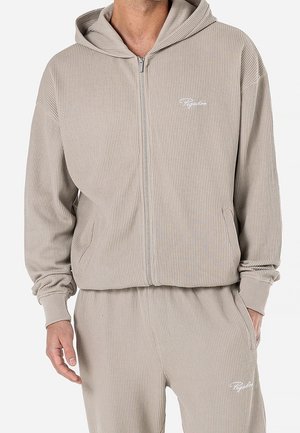 Hombre con sudadera con capucha beige de canalé con cremallera y pantalones deportivos a juego de canalé con logotipos en escritura blanca en el pecho izquierdo y el muslo.