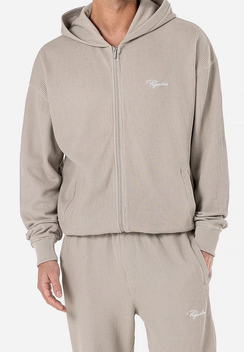 Homme portant un sweat à capuche zippé côtelé beige et un pantalon de jogging côtelé assorti avec des logos en écriture blanche sur la poitrine gauche et la cuisse.
