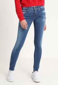 Jeans de denim azul ajustados con un lavado medio, ligeros desgastes y diseño de cinco bolsillos, combinados con zapatillas blancas y un sudadera roja.
