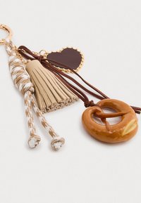 Porta-chaves com um encantamento de pretzel castanho, franjas em bege e um pendente de coração castanho escuro, com acabamentos em dourado e detalhes trançados.