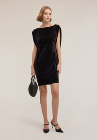 Schwarzes Samtkleid mit kurzen Ärmeln, glatter Textur, knielang, kombiniert mit einer schwarzen, mit Perlen verzierten Handtasche und spitzen Pumps.