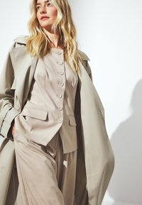 Bēša piegriezuma uzvalks ar pieguļošu žaketi, kurai ir seši pogas un kabatas, apvienots ar platu kāju biksēm, pārklāts ar vieglu tranču mēteli.