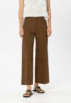 Frau trägt hoch taillierte, braune, weit geschnittene Hose, weißes ärmelloses Oberteil mit Blumendetails und schwarze flache Sandalen mit Schnallen.