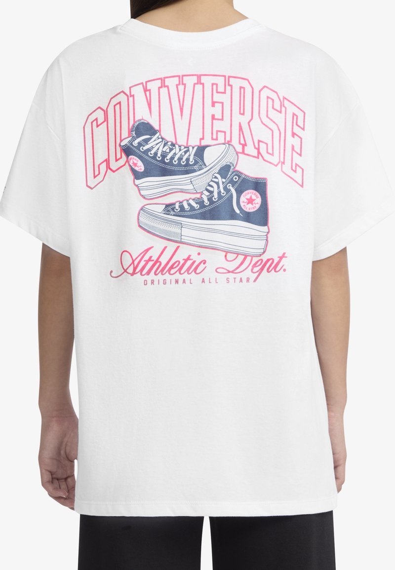 Personne portant un t-shirt blanc avec un logo rose "Converse" et un graphisme de baskets montantes bleues dans le dos.