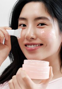 Maschera gel rosa in un barattolo trasparente etichettato "Abib." Una donna applica il gel con una spatola traslucida, mostrando una pelle liscia ed un'espressione concentrata.