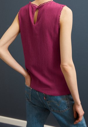Ærmeløs tekstureret magenta top med takkede kanter og nøglehulsryg båret med blå jeans mod en mørk baggrund.