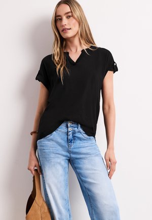 Blondynka nosząca czarną koszulę z podwiniętymi rękawami i jasnoniebieskie dżinsy, trzyma jasnobrązową zamszową torebkę przy boku na białym tle.