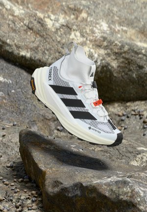 Chaussure de randonnée blanche Adidas Terrex Gore-Tex avec des bandes noires et des accents orange flottant au-dessus d'un terrain rocheux.
