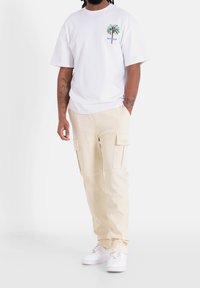 Vit t-shirt med grön palmlogga; beige cargobyxor med sidofickor; vita sneakers; medelbyggd, stående pose.