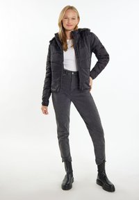 Jachetă puffer neagră cu glugă, design matlasat, fermoar frontal și buzunare laterale, purtată peste o cămașă albă și asortată cu blugi gri cu talie înaltă și botine negre.