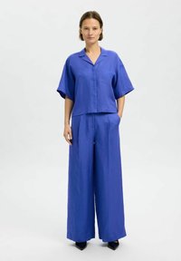 Mėlyna lininė trumpa marškinėliai su trumpomis rankovėmis ir kišene, derinami su plačiomis kelnėmis. Lygus paviršius, laisvas siluetas ir minimalistinis dizainas.