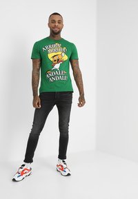 Vihreä puuvillapaita, jossa on sarjakuvahahmokuva, pyöreä kaula-aukko ja lyhyet hihat, yhdistettynä mustiin farkkuihin ja värikkäisiin lenkkareihin.